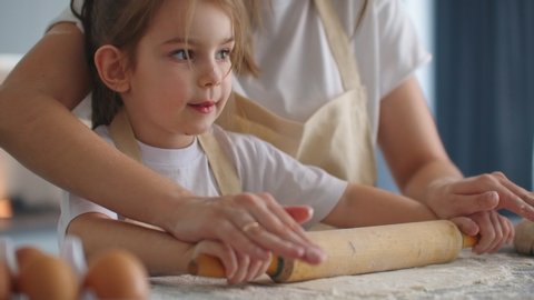 Mother Helping Daughter Roll Dough in Kitchen to Bake Cookies. Mom and daughter bake pizza in the kitchen together. Girl helps her mom to roll out the dough with a rolling pin. 