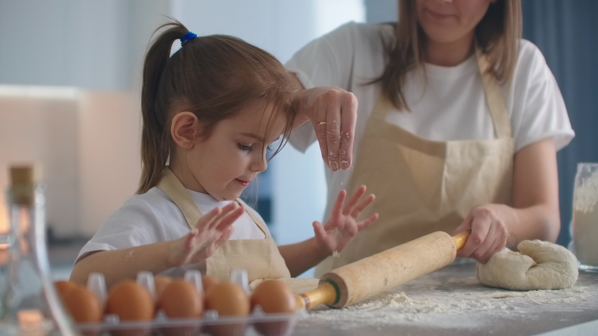Mother Helping Daughter Roll Dough in Kitchen to Bake Cookies. Mom and daughter bake pizza in the kitchen together. Girl helps her mom to roll out the dough with a rolling pin.  Royalty-Free Stock Footage #1048572586