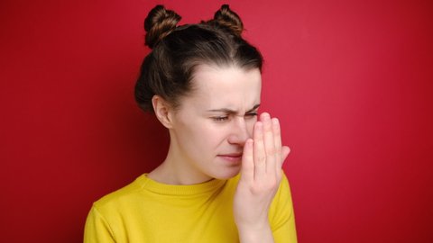 Unhappy young woman checking her breath with hand, smells something awful, pinches nose, frowns in displeasure, dressed in yellow sweater, isolated over red background. Bad smell concept.