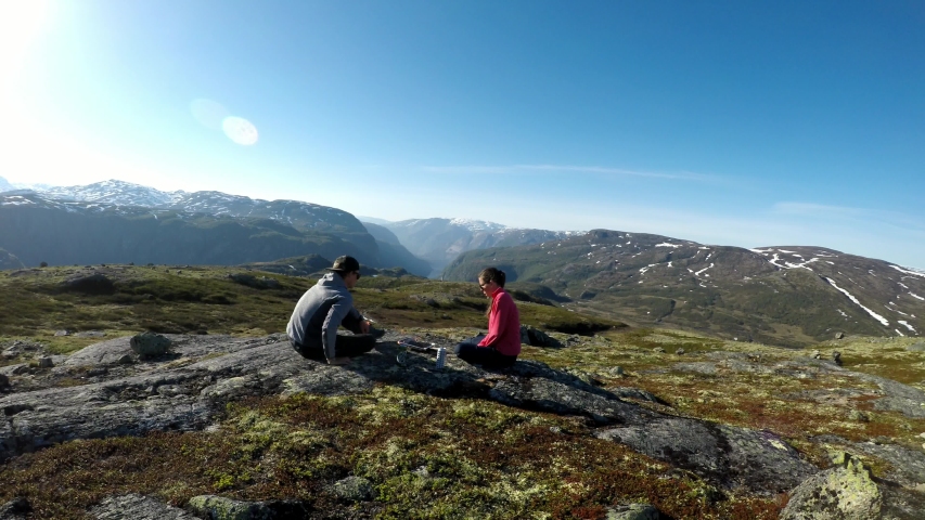 A couple in hiking outfit camping in the wilderness, at the top of fjord with the view on Eidfjord. They are preparing hot dogs on the grill and sipping a beer. Being close to nature. Having fun

