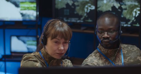 Multiethnic man and woman, military dispatchers colleagues in headsets sitting in monitoring room with screens and controlling troops operation. Two mixed-races officers cooperating in control room.