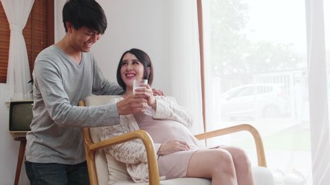 Asia new couple family live in house. Pregnant woman sit on sofa hold glass of milk, husband give milk then girl drink milk and look at him with happy smile face at home. Baby family pregnancy concept