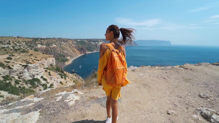 super slow motion Rear view of taned traveler woman standing on cliff edge in front of amazing seascape,posing in bright yellow dress blowing in the wind. Freedom, travel and summer vacation.