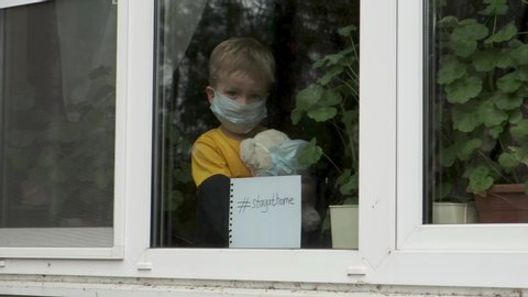 Stay at home quarantine coronavirus pandemic prevention. Sick child with teddy bear in protective medical masks sits on windowsill and looks out window. View from street. Prevention epidemic.