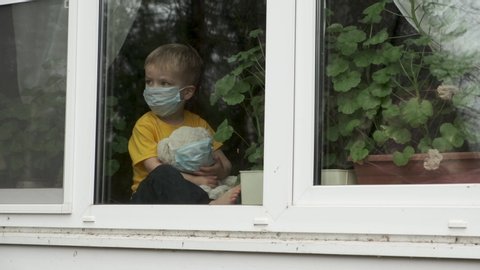 Bored child on home quarantine. Boy with teddy bear both in protective medical masks sits on windowsill and looks out window. Allergy, covid-19 coronavirus pandemic, prevention epidemic.