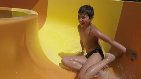 Funny laughing boy is rotating and riding down orange slide in a water park. Boy in a swimsuit laughs and slides into the water raising spray