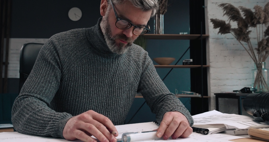 Attractive Architect working at his Workplace with pencil and Ruler, making Drawings. Having blueprints on the Desk. Looking successful and Serious, working with papers and Calculations.
