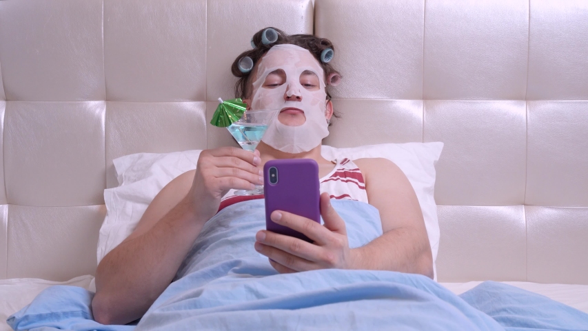 Strange man with a cosmetic mask on his face and curlers does spa treatments at home, holding a cocktail, takes selfie photo for social networks. Parody on cosmetics beauty and fashion bloggers | Shutterstock HD Video #1048617976