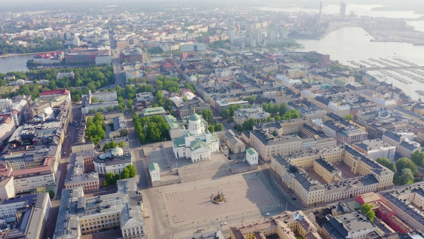 Helsinki, Finland. Helsinki Cathedral dominates the northern edge of Senate Square, its white neoclassical facade and tall green central dome visible from an elevated aerial perspective that sweeps across the historic city center. The cathedral was designed by Carl Ludvig Engel in a Greek cross plan with Corinthian columns and four smaller corner domes, completed in 1852 after more than three decades of construction. The surrounding Empire-style ensemble includes uniform yellow and white buildings, a spacious cobblestone plaza, and tree-lined streets that extend toward the harbor and Baltic Sea. The scene is captured in bright summer daylight with clear skies, the camera orbiting and pulling back to reveal the geometric grid of the old town, green parks, and the waterfront. The composition emphasizes the cathedral