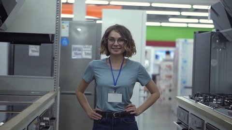 Positive female seller or shop assistant portrait in supermarket store. Woman in blue shirt and empty badge looking at the camera and smiling. Household appliances on the background. Close up camera