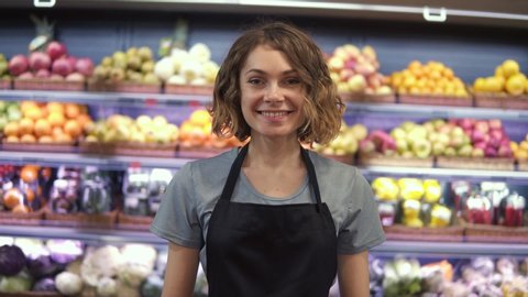 Portrait of attractive young saleswoman in black apron standing in supermarket with shelves of fruits on background, looking at camera and smiling. Trade business and people concept
