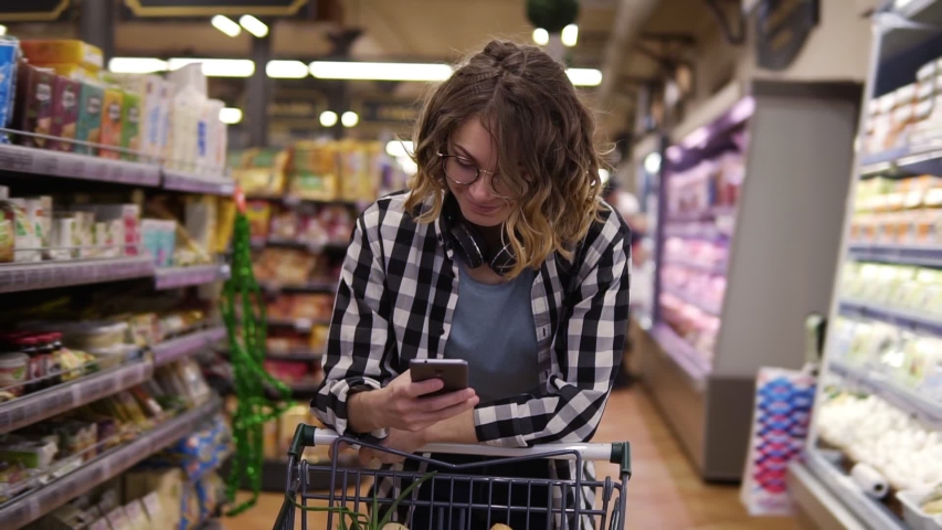 Shopping, technology, sale, consumerism and people concept - woman with smartphone and headphones on neck walking at supermarket. Smiling woman texting with friends, leaning on her shopping trolley