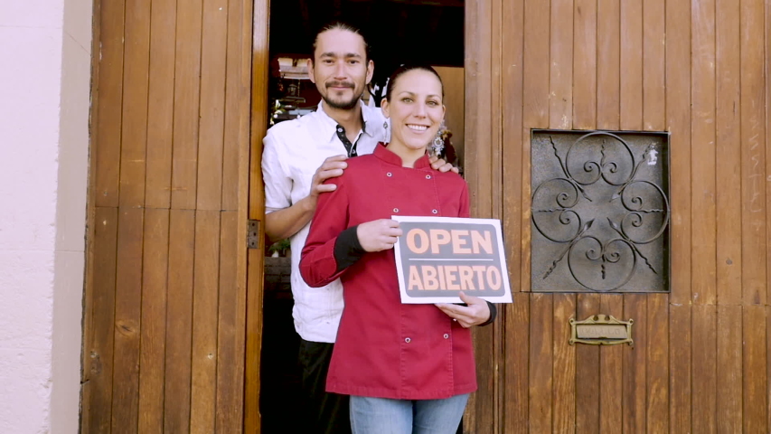 Young, attractive, happy, smiling, Hispanic couple in their 30s holding a bilingual open sign in front of their business in slow motion Royalty-Free Stock Footage #1048623946
