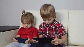 Children in red shirts are lying on a bright sofa playing on a tablet in a social network. Blond boy and girl watch videos on a digital tablet.