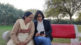 Lifestyle video shot of a young Indian woman sitting with her mother in law in a park. Older senior woman reads a joke from the phone and both of  them share a laugh. Concept - Digital literacy 