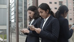 Focused businesswomen using smartphones. Concentrated female colleagues using mobile phones while standing outside urban building. Technology concept