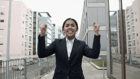 Excited young businesswoman celebrating success. Front view of cheerful young businesswoman raising fists and showing thumbs up on city street. Winner concept 