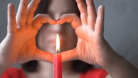 A young woman is sitting at a table. Burning candle Hands in the form of a heart. Symbol of love. St. Valentine's Day. Holiday. Declaration of love. Close-up.