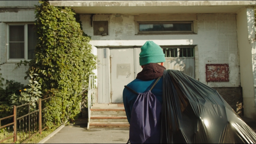 young man in green knitted hat with huge plastic bag of garbage comes to dwelling building entrance backside view