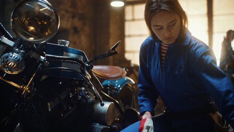 Young Beautiful Female Mechanic is Fixing a Custom Bobber Motorcycle. Talented Girl Wearing a Blue Jumpsuit. She Uses a Ratchet Spanner and Checks Tablet for Advice. Creative Authentic Workshop Garage
