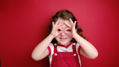 Cheerful funny little curly girl having fun making glasses shape with hands, does funny face with eyes wide open, dressed in white t-shirt, isolated on red background. People emotions concept