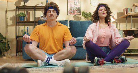 Funny retro style couple of Caucasian man and woman meditating on yoga mat indoors. Pretty smiling happy young girl fooling around with man while practicing meditation at home. Sport concept