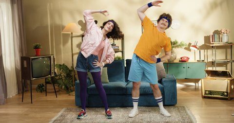 Cheerful Caucasian male and female retro style models stretching muscles while standing in room. Funny couple of man and woman exercising at home near sofa. Indoor pair workout at home. 50s concept