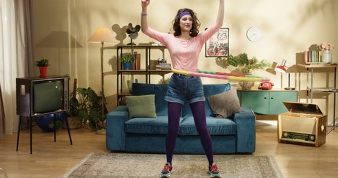 Pretty Caucasian retro style female model in purple tights doing exercise with hula hoop near blue couch at home. Beautiful stylish funny girl working out with equipment in living room. 70s concept