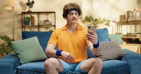 Retro style Caucasian funny sportsman video chatting on smartphone and showing up his muscles while sitting on blue sofa at home. American style athlete of 50s showing biceps to phone camera indoors