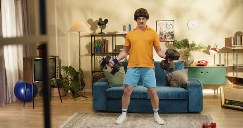 Strong funny Caucasian athlete with mustache standing in room and working out with big dumbbells. Retro style sportsman doing exercise on biceps with equipment while having home training. 50s concept