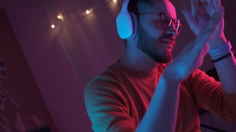 Handsome man listening music using headphones and mobile phone in living room in colourful neon light. Student resting after work and enjoying music. Dance smooth hand movements to the rhythm