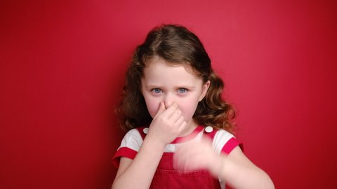 Unhappy little girl covers nose with hand, smells something awful, pinches nose, frowns in displeasure, dressed in white t-shirt, isolated over red background. Smelling bad gesture and odor concept.