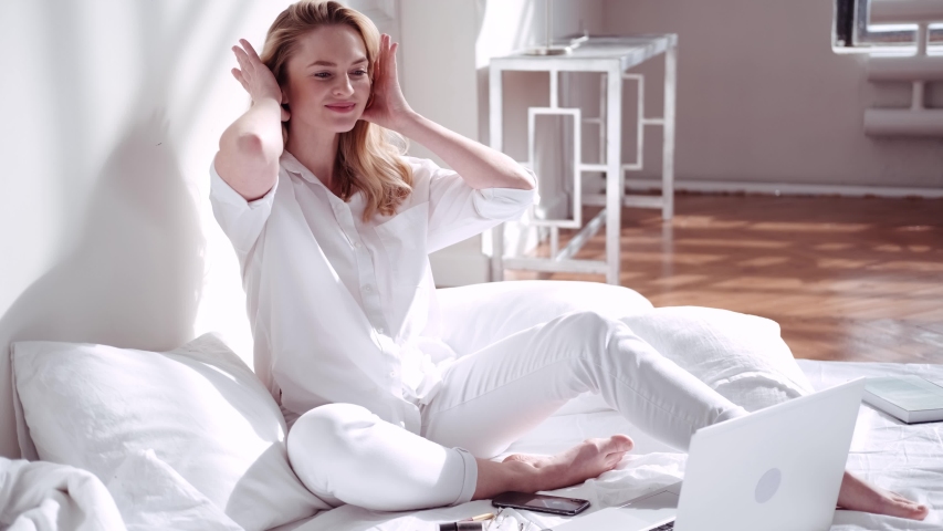 A beautiful woman in white pajamas collects her hair and does a careless hairstyle (bun), staying in bed on a sunny morning. Day off, bright interior of the house or hotel. Special day. Royalty-Free Stock Footage #1048681945