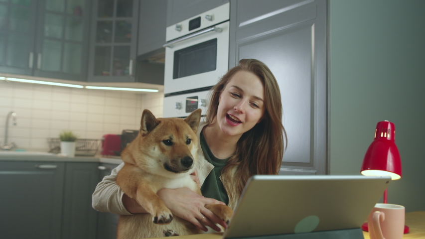 Side View of Young Woman Sitting at Table and Petting Her Dog While Making Video Call. Attractive Girl Video Chatting, Smiling, Laughing, Talking. Online Communication Concept. Slow Motion Shot