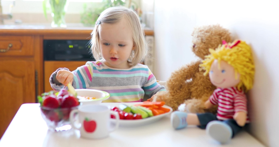 Adorable toddler girl eating fresh fruits and vegetables for lunch. Child feeding doll and teddy bear in the kitchen. Delicious healthy food for kids Royalty-Free Stock Footage #1048684741