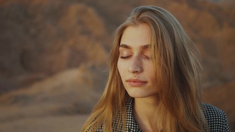 Portrait of pretty blond woman with closed eyes on sunset. Happy young woman standing in a desert mountains, sunset landscape, slow motion, 4k