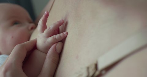 Mom caresses her baby's little palm at chest during breastfeeding in room. Slowly in 4K