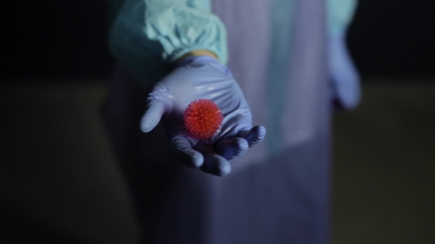 Close up of a scientist in protective medical clothing, suit pointing to the camera a coronavirus COVID-19 model. Dark background.