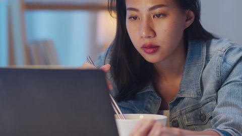 Close up shot : Asian freelance business women eating instant noodles and working on computer laptop in living room at home at night. Hungry young Asia girl sitting and work at home concept.