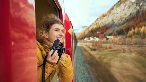 Excited happy woman, smiling and laughing leans out of train carriage window. Traveling young woman with photocamera during amazing trip on bernina express train
