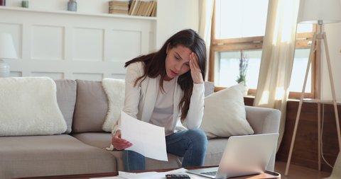 Upset young woman feeling stressed about financial debt checking bills at home. Frustrated lady doing paperwork sitting on sofa thinking of unpaid tax, bank loan, money problem or bankruptcy concept.