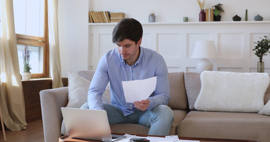 Young man checking household bills to pay online at home. Millennial guy holding paper bank statement calculating tax, managing finances and expenses, bank loan payment sitting on sofa in living room. Royalty-Free Stock Footage #1048713307