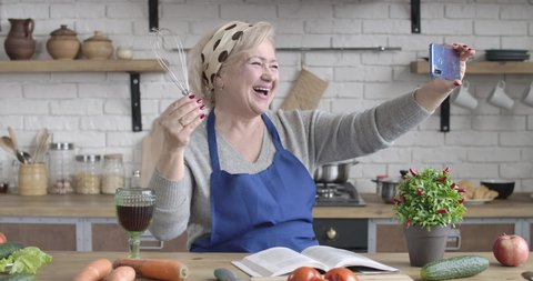 Portrait of laughing senior Caucasian woman posing with whisk and wineglass at selfie camera. Mature positive retiree having video call on smartphone. Cinema 4k ProRes HQ.
