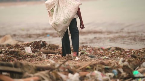 Man is walking along the worlds most polluted beach in Bali. Plastic bags bottles and paper trash on the sandy dirty beach. Sea waves full of litter hitting the beach. Huge dump in tropical paradise.
