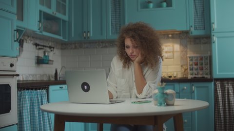 Curly 20s woman tired of working at laptop on home kitchen. Female freelance home worker closing laptop to have a break. Sad working woman sitting at the table. 