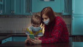 30s mother and boy in medical masks talking online by mobile phone with relatives, greeting waving hands. Family at home in self isolation from virus. Stop epidemic. 