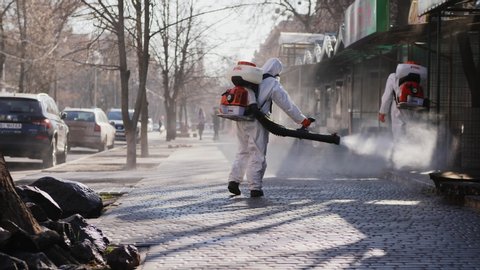 POLTAVA, UKRAINE - CIRCA March 2020: People wearing protective clothing for spraying chemicals disinfect the sidewalk and buildings on the street to prevent the spread of the corona virus