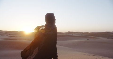 Beautiful girl in traditional Abaya dress and in hijab enjoys nature in the desert at sunset. 4K Slow Motion