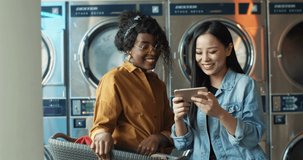 Multiethnic stylish young girls talking and watching photos or video on smartphone. Friends standing in laundry service. African American and Asian women with phone while washing machines working.