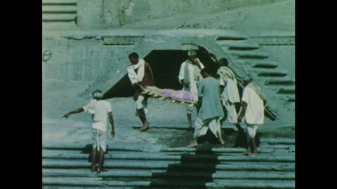 1960s: Men carry wrapped corpse down stone steps. Men arrange logs on stone slab.
